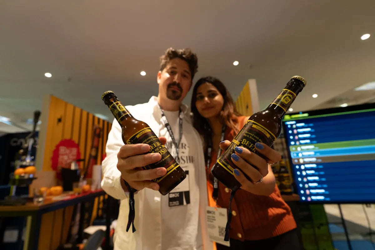 Two Beer of Satoshi hosts offering Genesis Lager bottles toward the camera